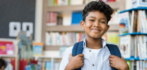 Portrait of smiling hispanic boy looking at camera. Young elementary schoolboy carrying backpack and standing in library at school. Cheerful middle eastern child standing with library background.